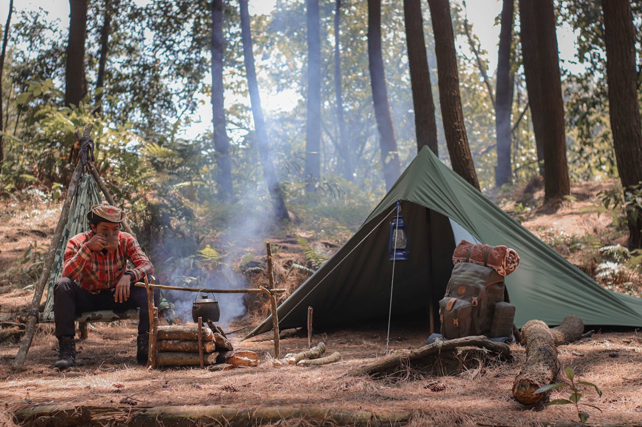 Man camping in forest, sitting by campfire with tent and backpack.